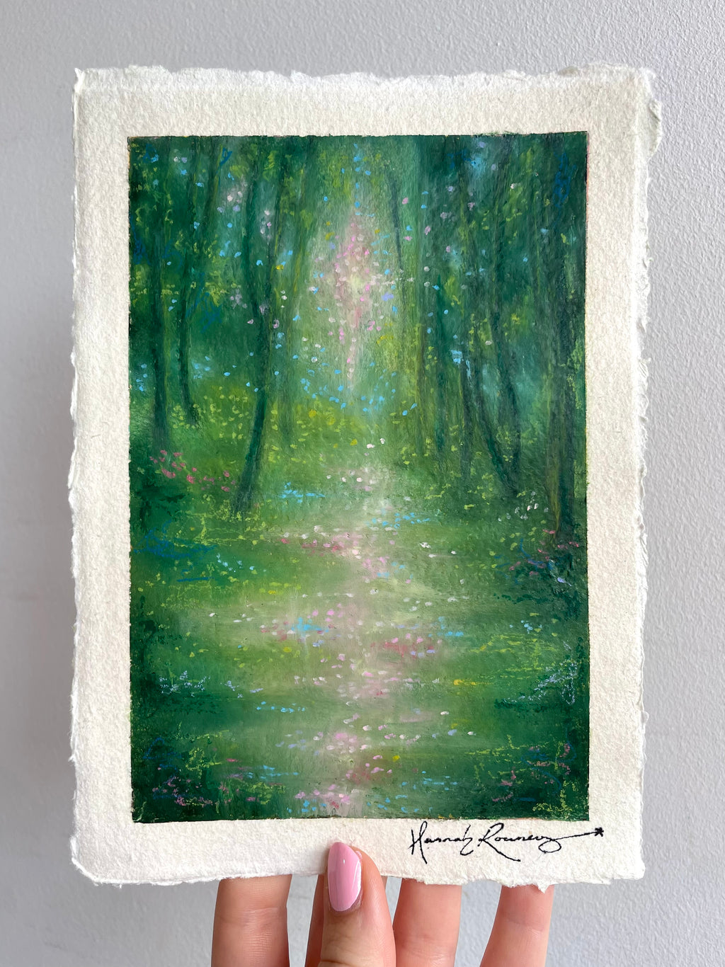 A hand holds an original painting on handmade paper, featuring a painted green forest with light filtering through the trees. The print has a textured off-white border and a visible artist signature at the bottom right.