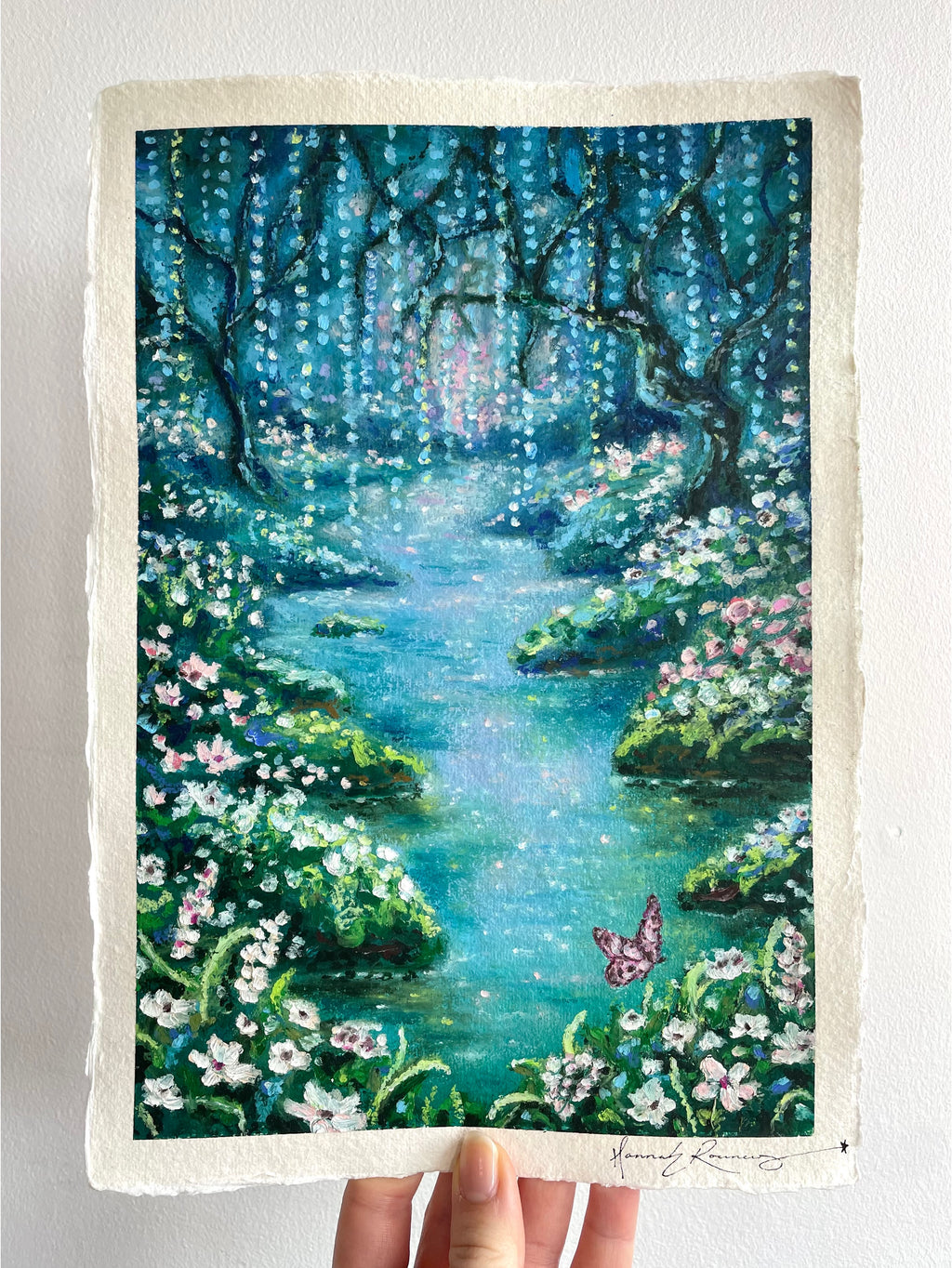 Whimsical oil pastel enchanted forest painting held in hand, featuring a blue-toned river, wildflowers, butterfly, and weeping willow against a white background.