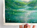 A close-up of a textured oil pastel painting of a green meadow and glowing river with abstract wildflowers, held by a hand against a white background.