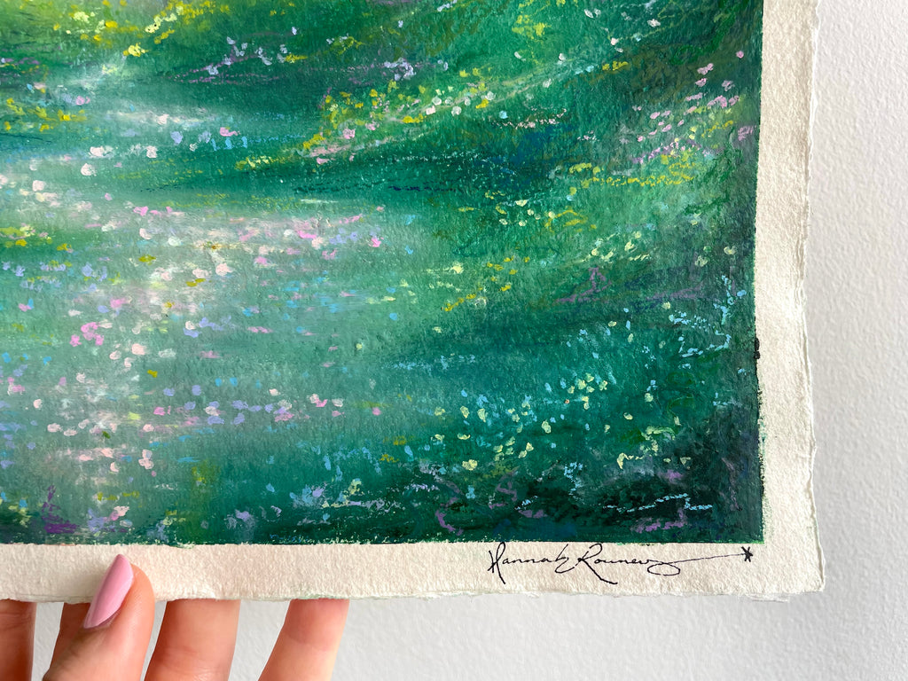 A close-up of a textured oil pastel painting held by a hand against a white wall. The artwork features a vibrant green and teal landscape with splashes of yellow, purple, and white.